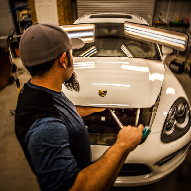 Person with a cap and beard working on the open hood of a white Porsche in a garage.