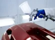 A gloved hand using a professional paint spray gun to paint a car bumper glossy red in a spray booth.